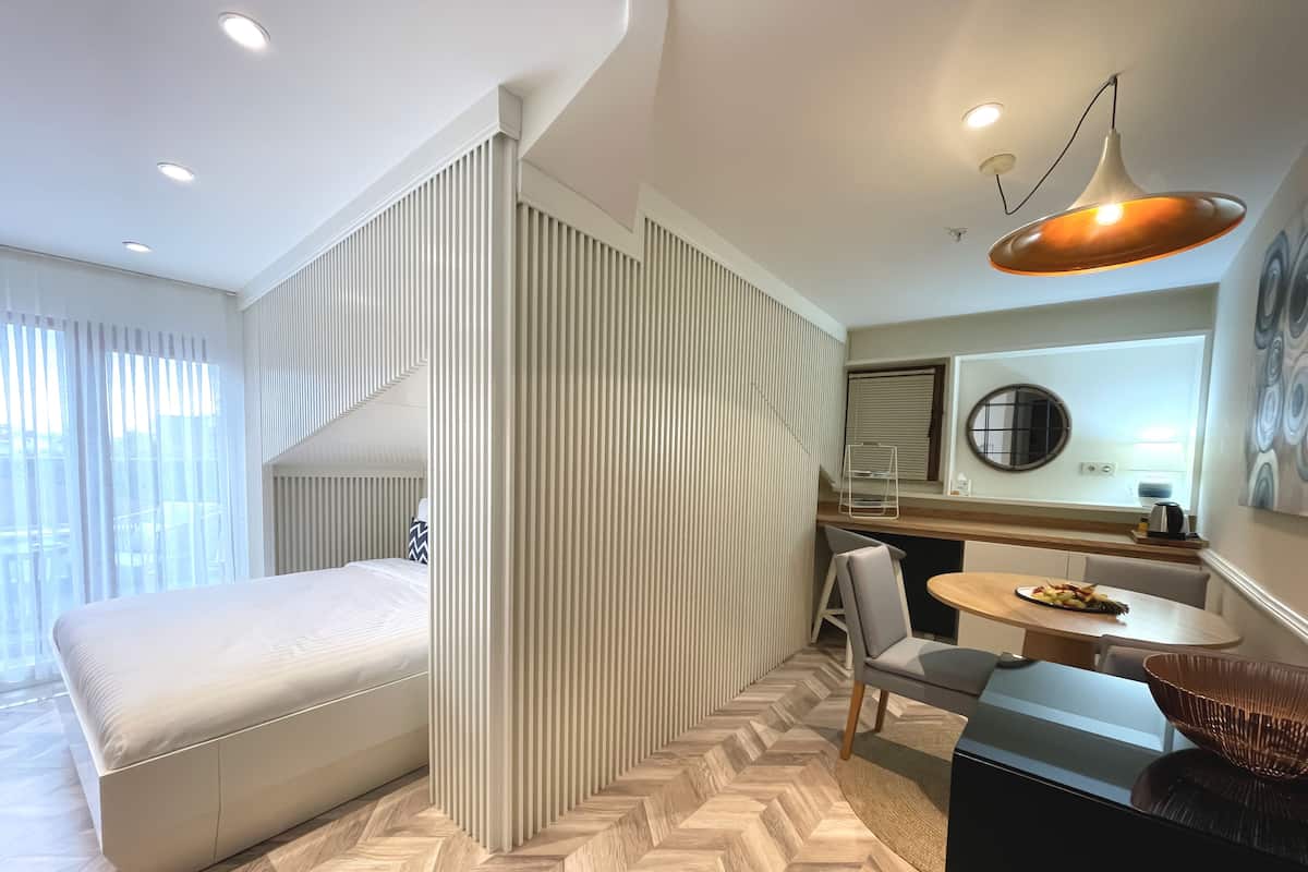 A modern hotel room with a bed, a dining area with a table and chairs, a kitchen counter, and a large window with curtains.