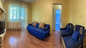 Uma sala de estar com um sofá azul, uma televisão sobre um móvel e piso de madeira.