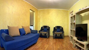 Uma sala de estar com um sofá azul, duas poltronas, uma TV e piso de madeira.