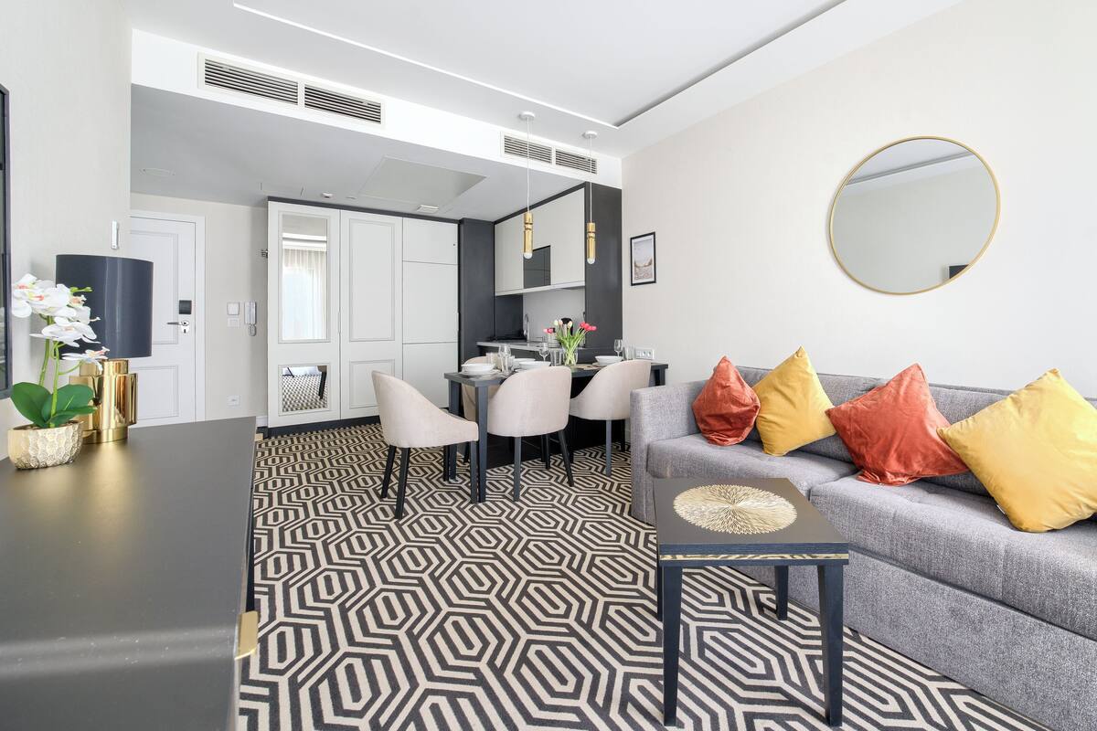 Een moderne woonkamer met een grijze bank, een eettafel met stoelen, een keuken op de achtergrond en een vloer met patroon.