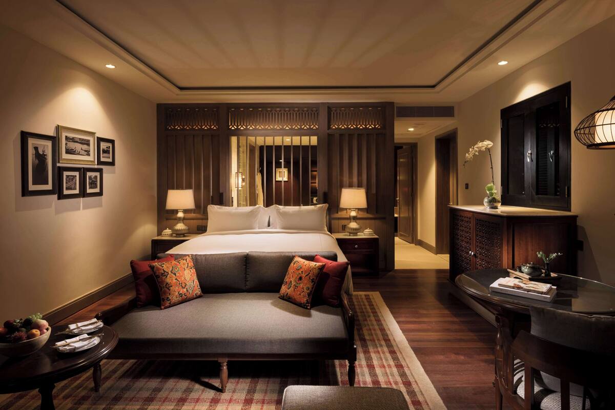 A hotel room with a large bed, two bedside lamps, a small table with a bowl of fruit, and a desk with a chair.