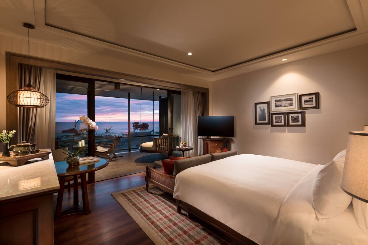 A hotel room with a large bed, a TV, a dining area with a table and chairs, and a balcony with a view of the sunset.
