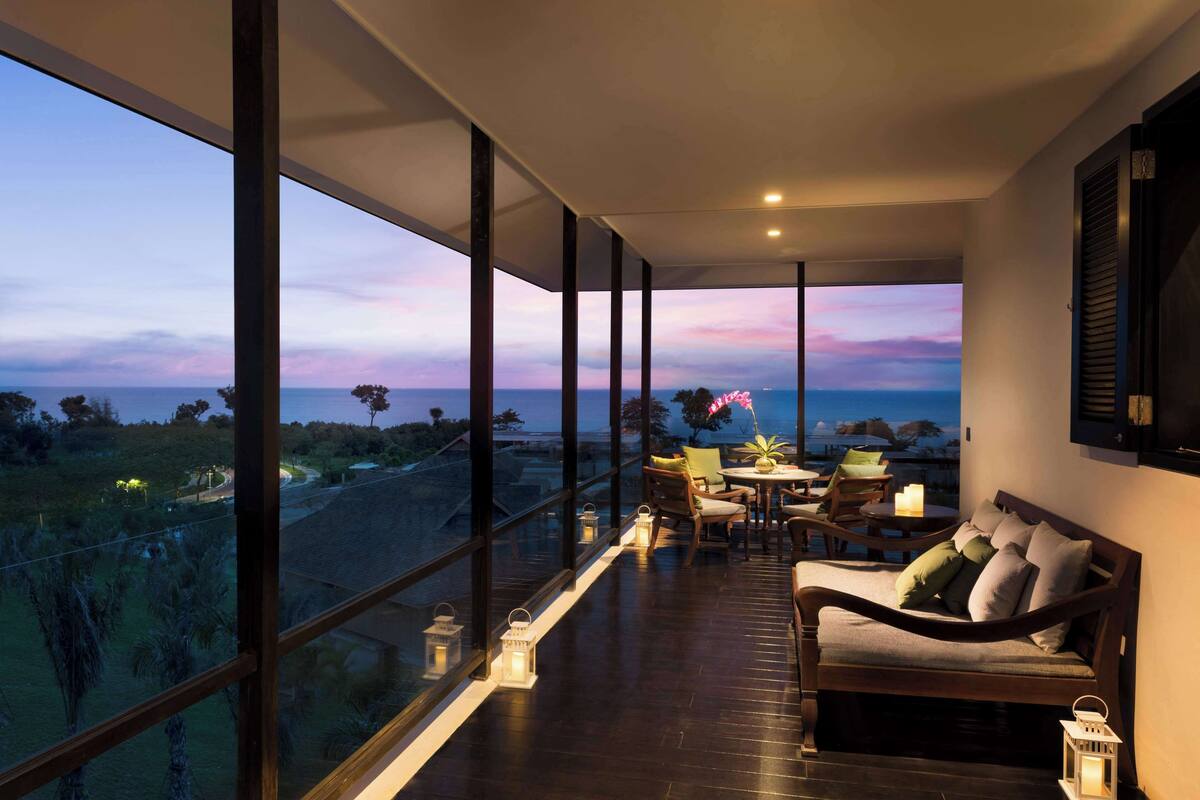 A balcony with a wooden floor, a sofa, a table with chairs, and a view of the landscape.