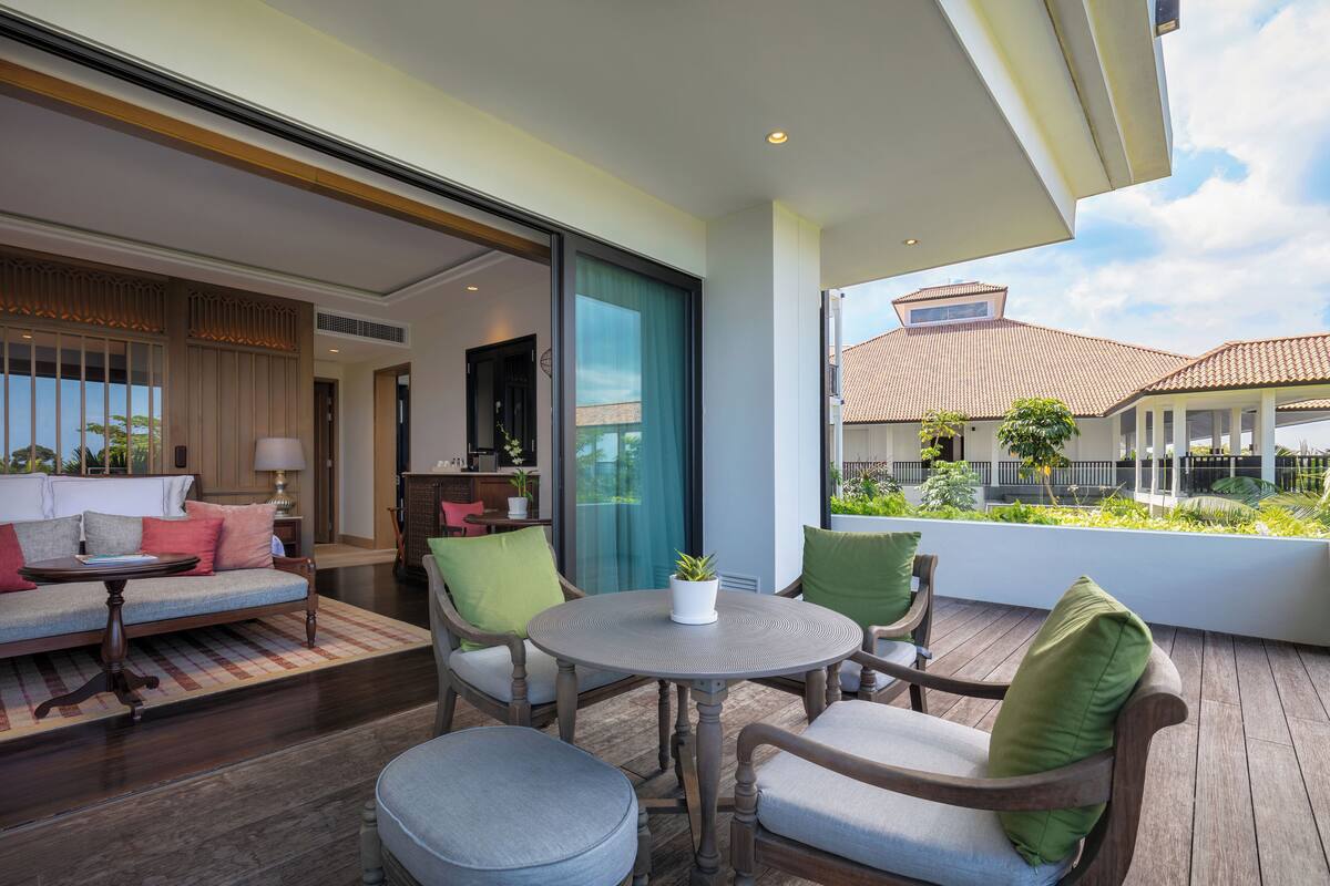 A modern living room with a sofa, chairs, and a round table. There is a balcony with a view of a building and greenery.