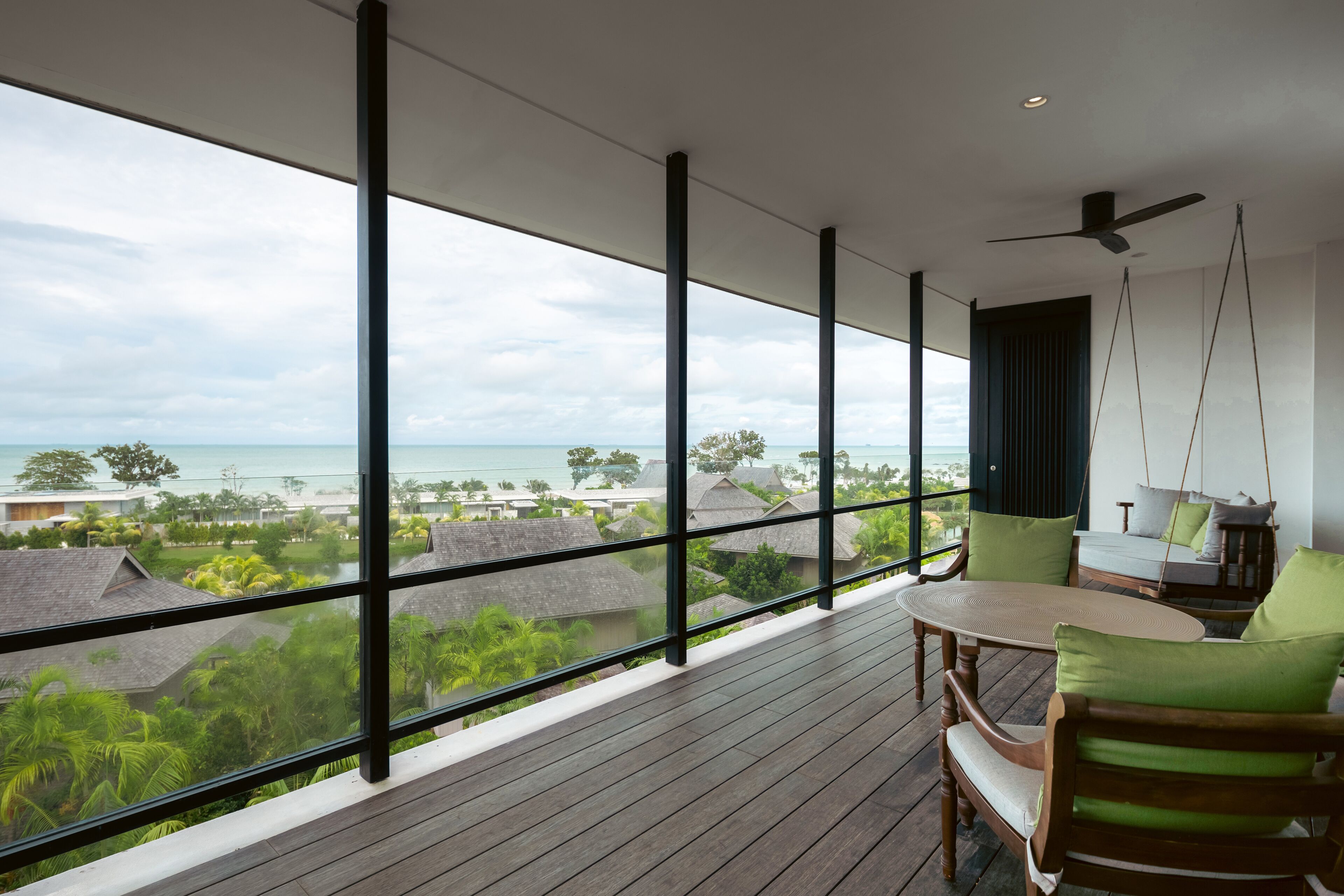 Ein Balkon mit Blick auf das Meer und Häuser, Holzmöbel und ein Deckenventilator.