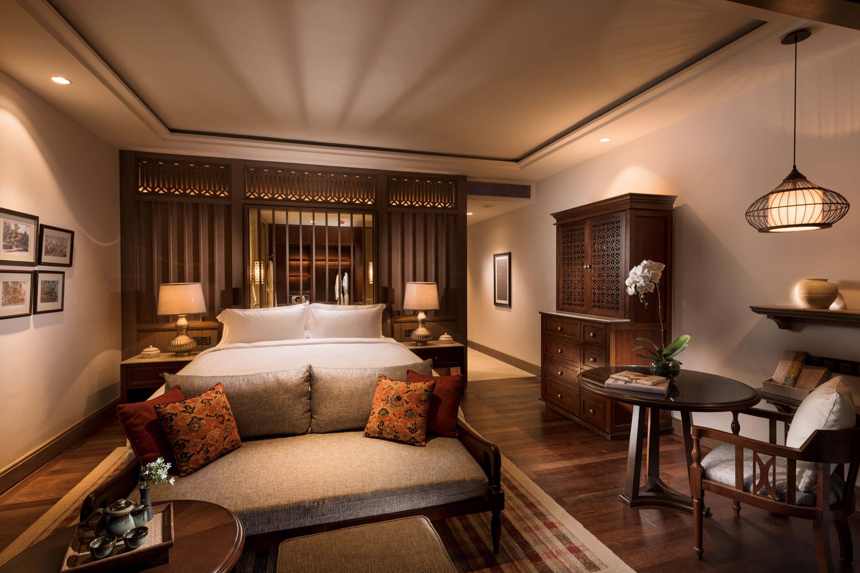 Een hotelkamer met een groot bed, twee nachtkastjes met lampen, een houten kast, een ronde tafel met stoel, en een decoratieve hanglamp.