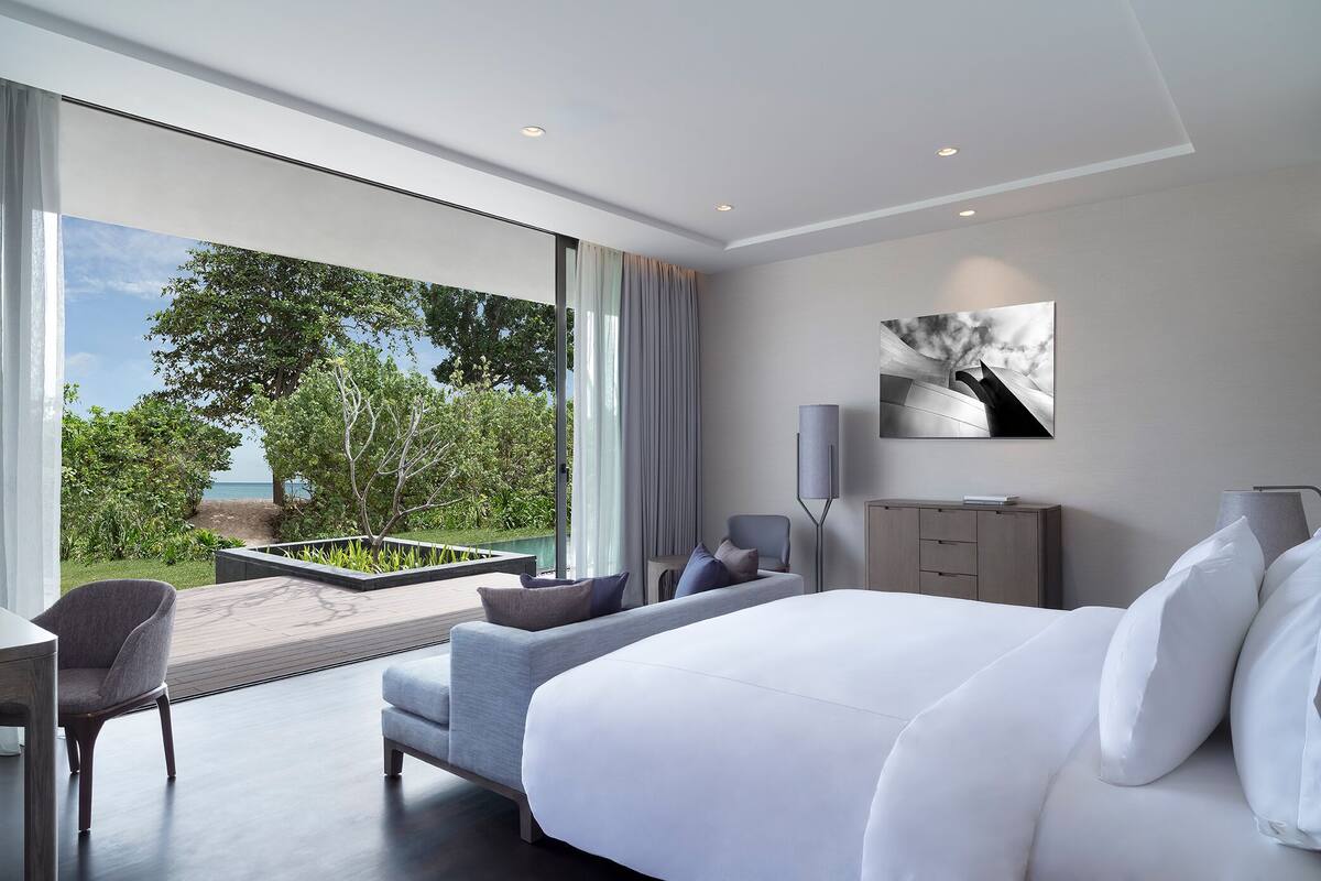 A modern hotel room with a large bed, a seating area with a sofa and armchairs, a desk, and a view of greenery and the sea through large windows.