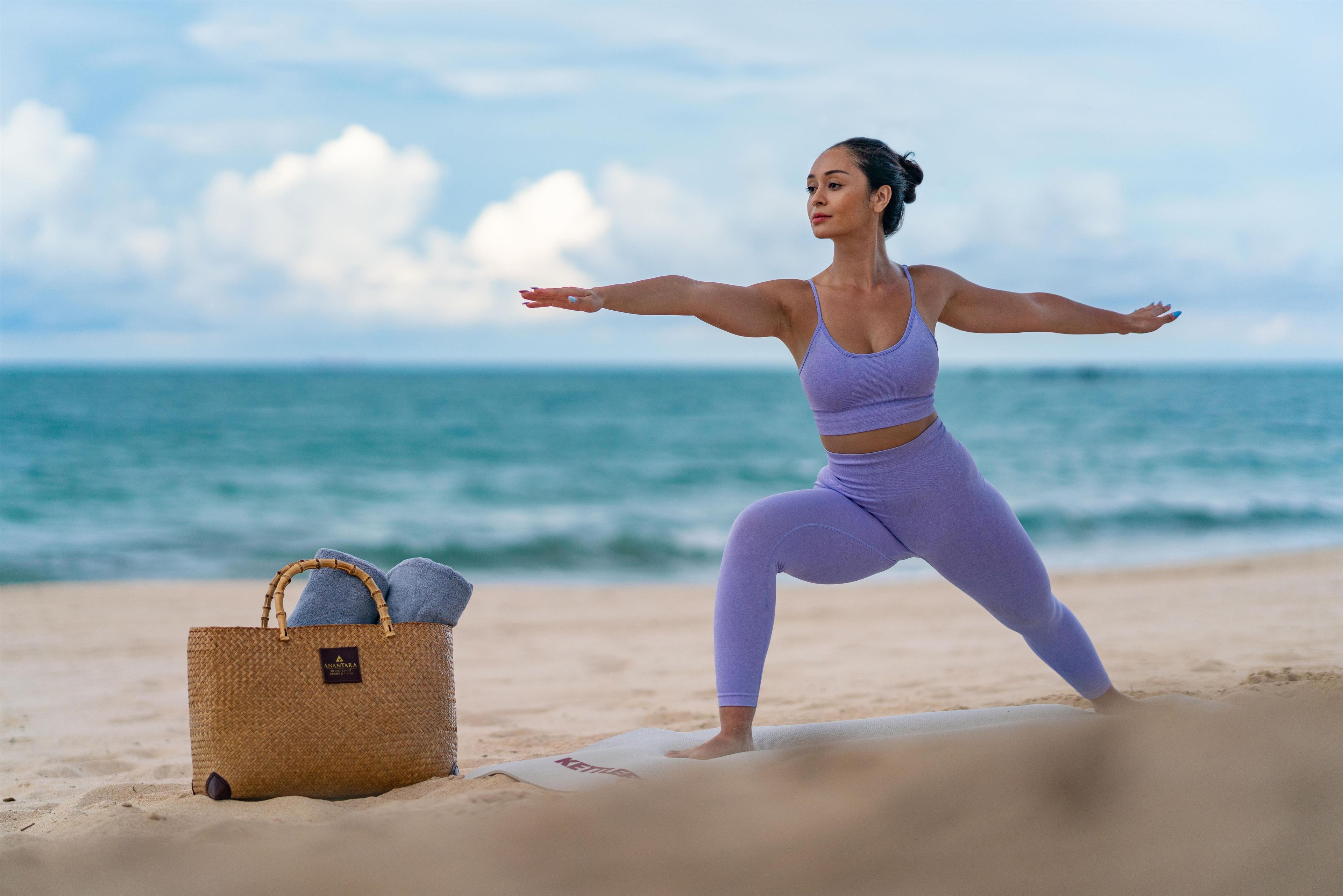 Une personne pratiquant le yoga sur une plage, avec un panier rempli de serviettes et un tapis.
