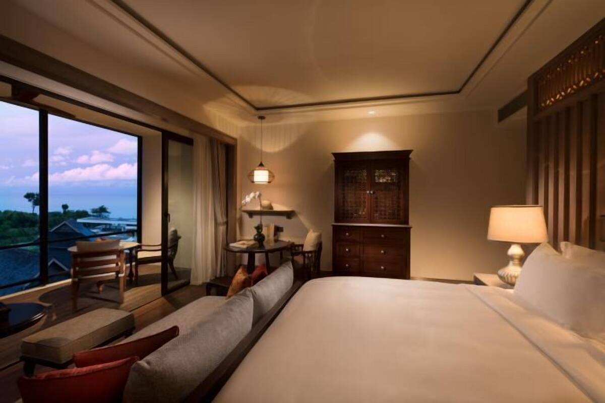 A hotel room with a large bed, a desk with a lamp, a chair, a small table, and a view of the ocean.