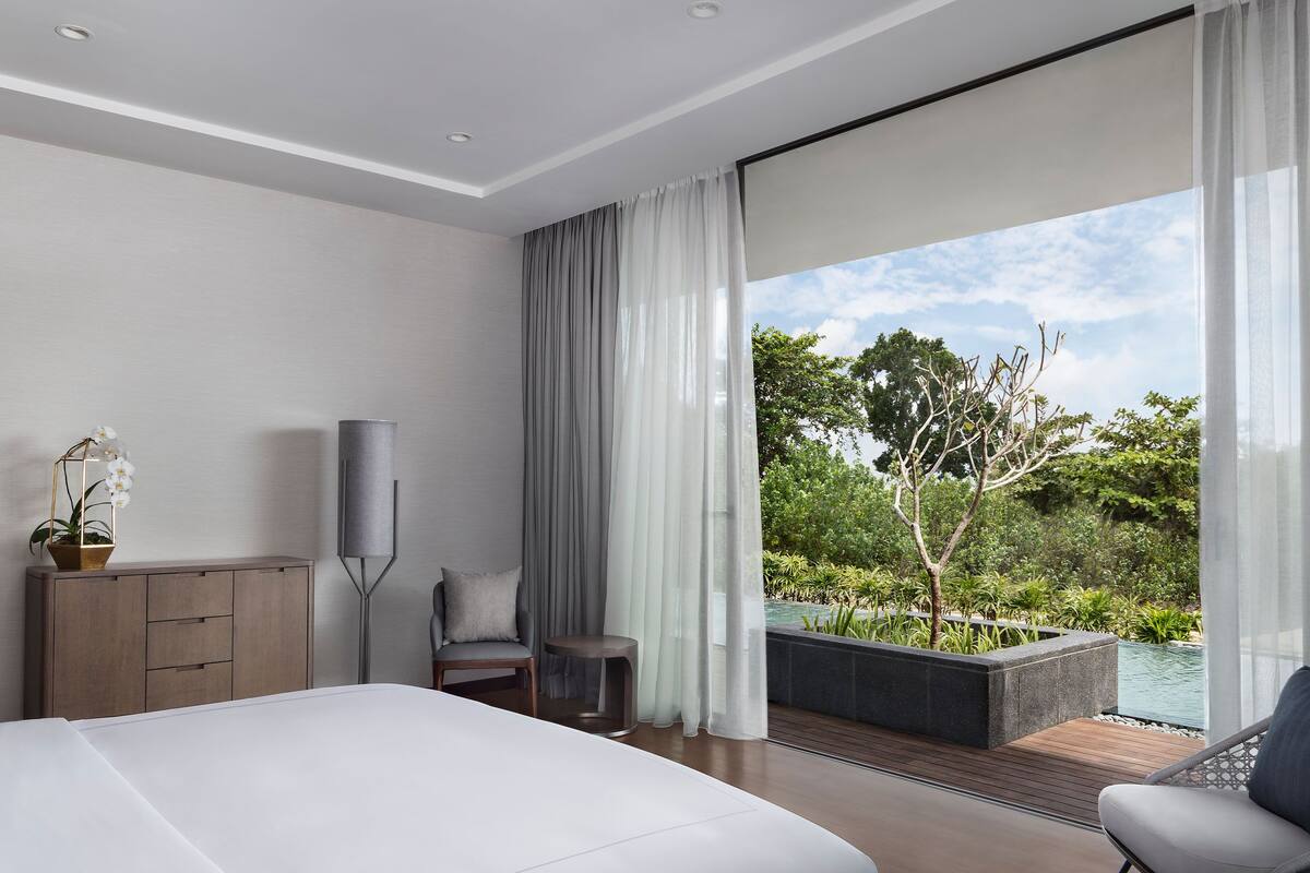 A modern bedroom with a large bed, a TV, a chair, and a view of a garden and pool.