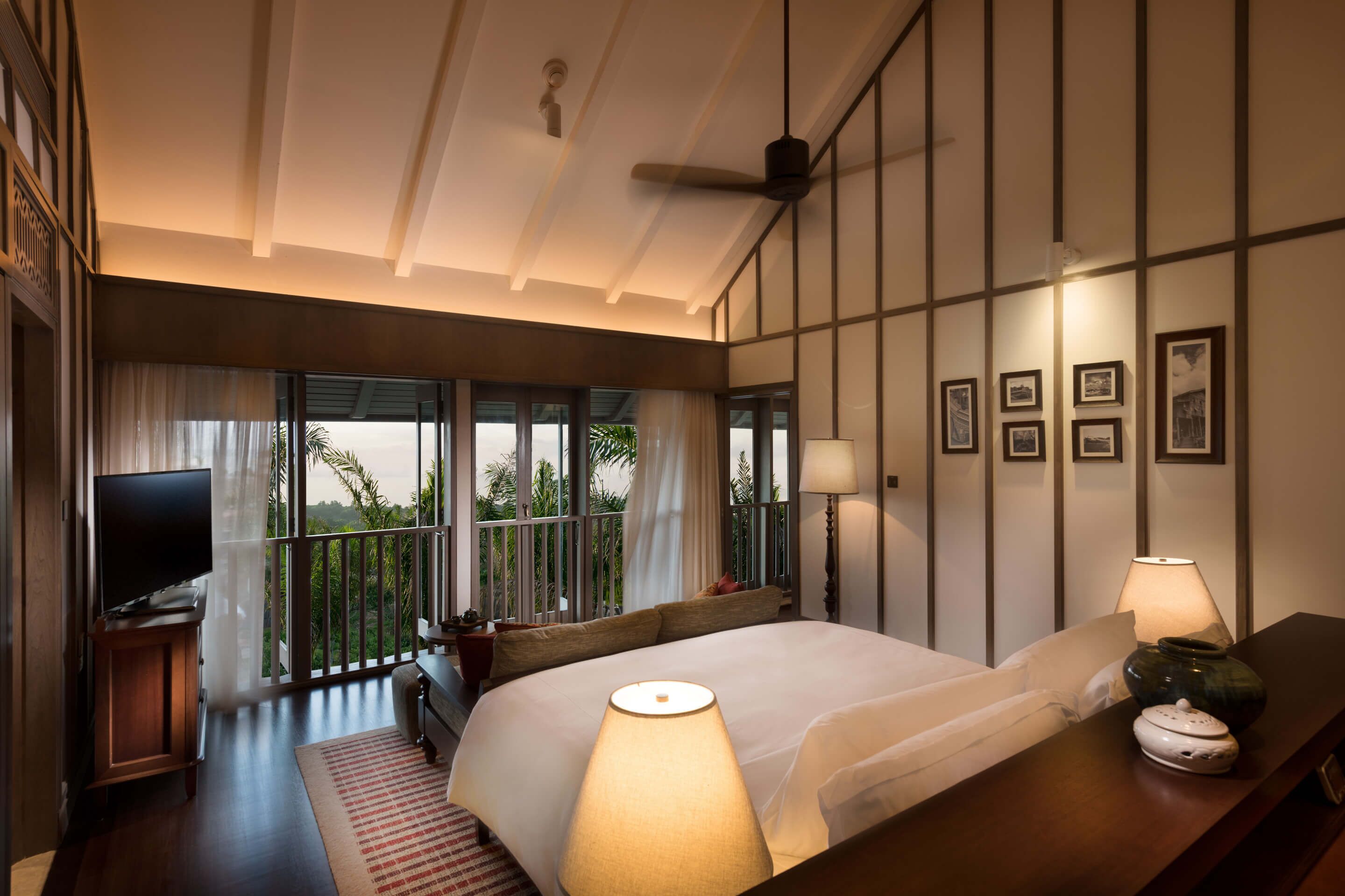 A hotel room with a large bed, a TV on a wooden stand, a balcony with curtains, and framed pictures on the wall.