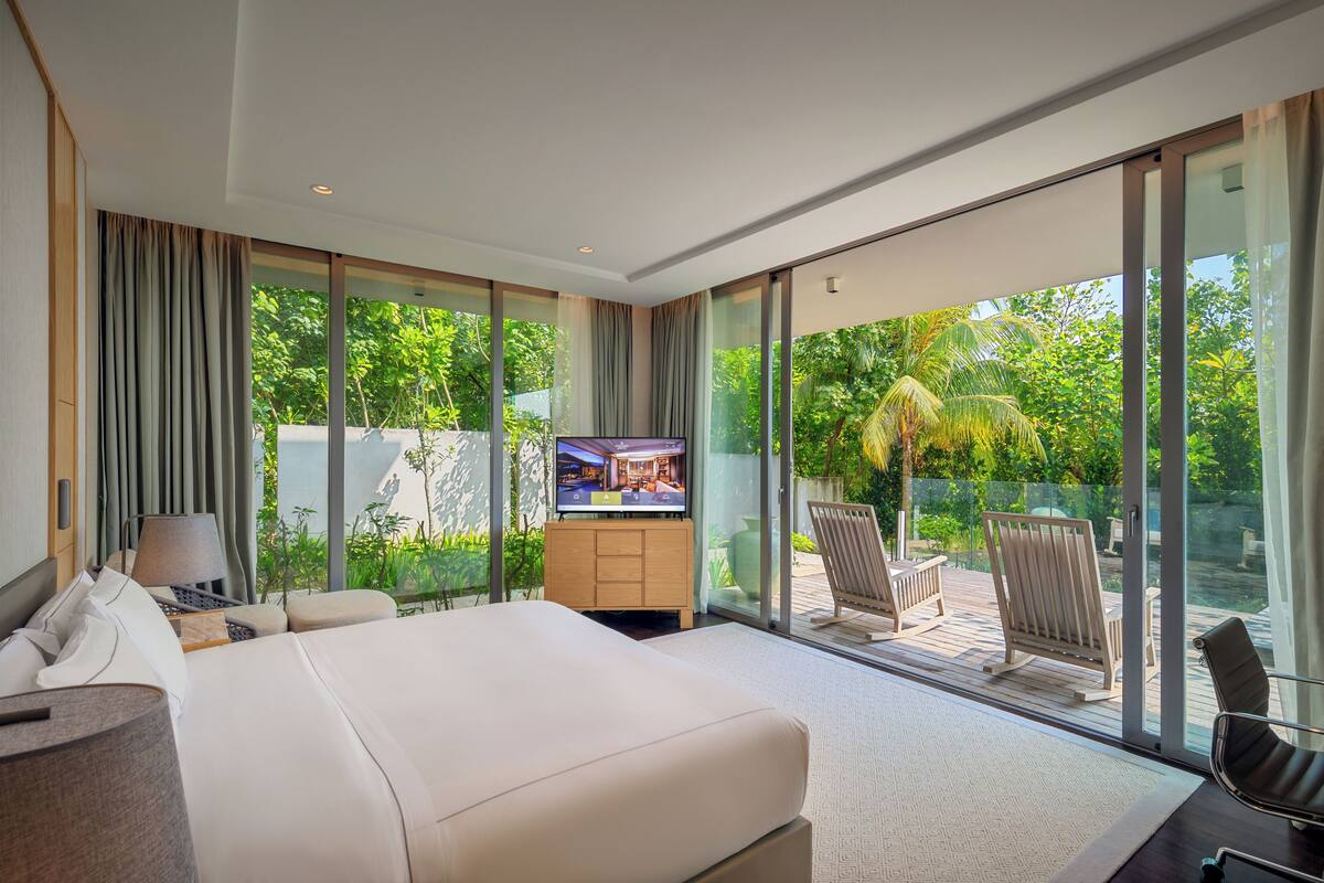 A modern hotel room with a large bed, a television, a desk, and a balcony with outdoor seating.