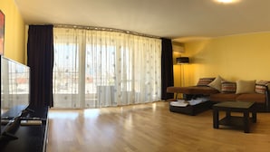 A living room with a sofa, a coffee table, a TV, and a large window with curtains. - Apart holiday Coralia Mamaia CityLiving (Constanta)