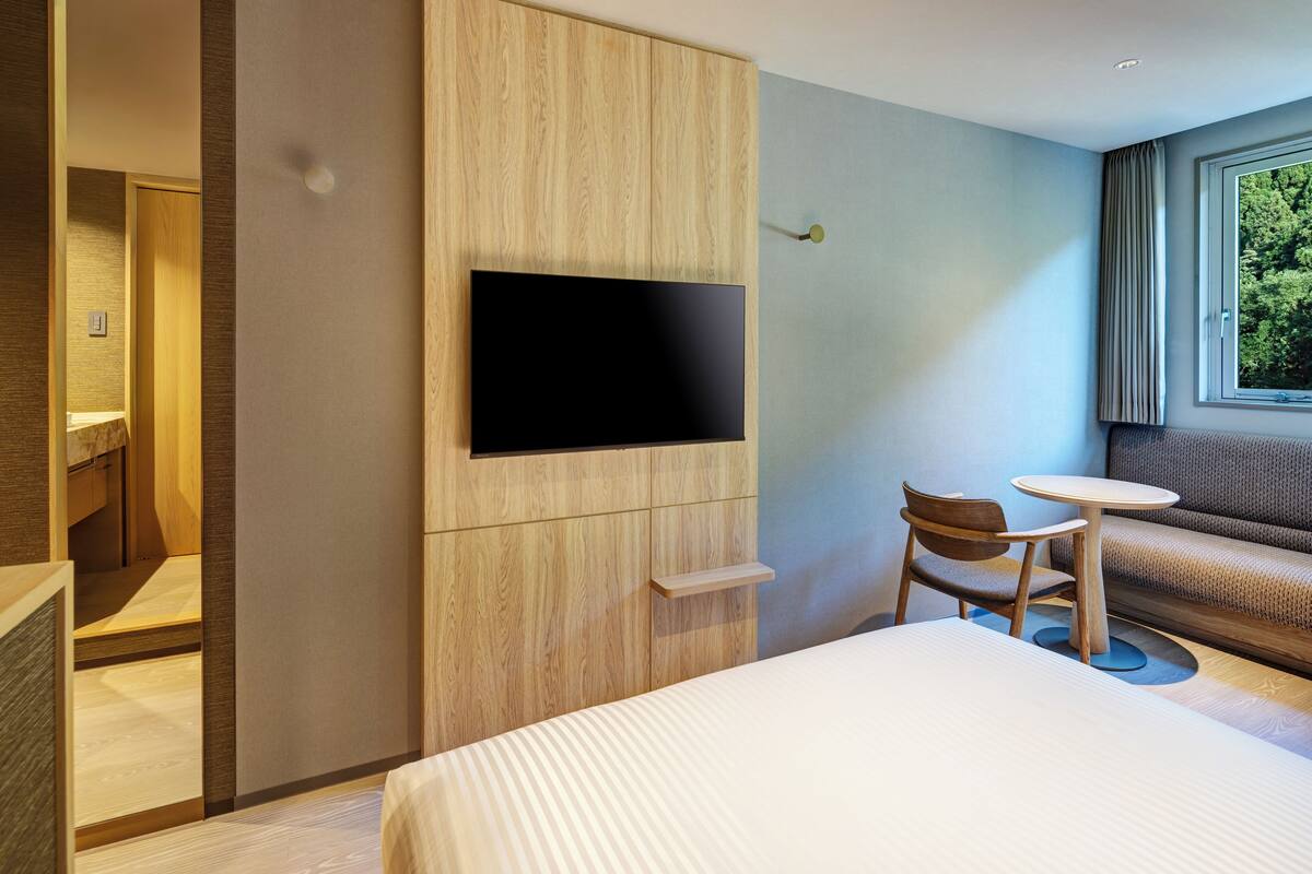 A modern hotel room with a bed, a TV mounted on a wooden panel, a small round table, and a sofa.