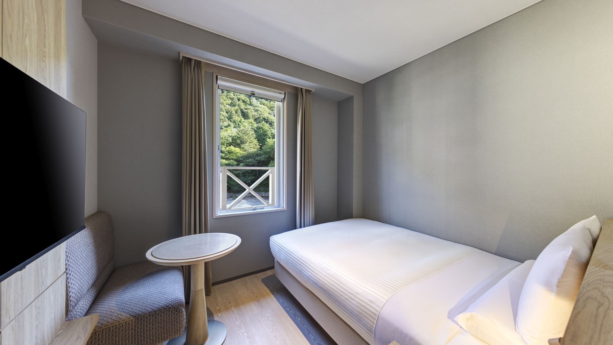 A hotel room with a bed, a TV, a small table, and a window with a view of trees.