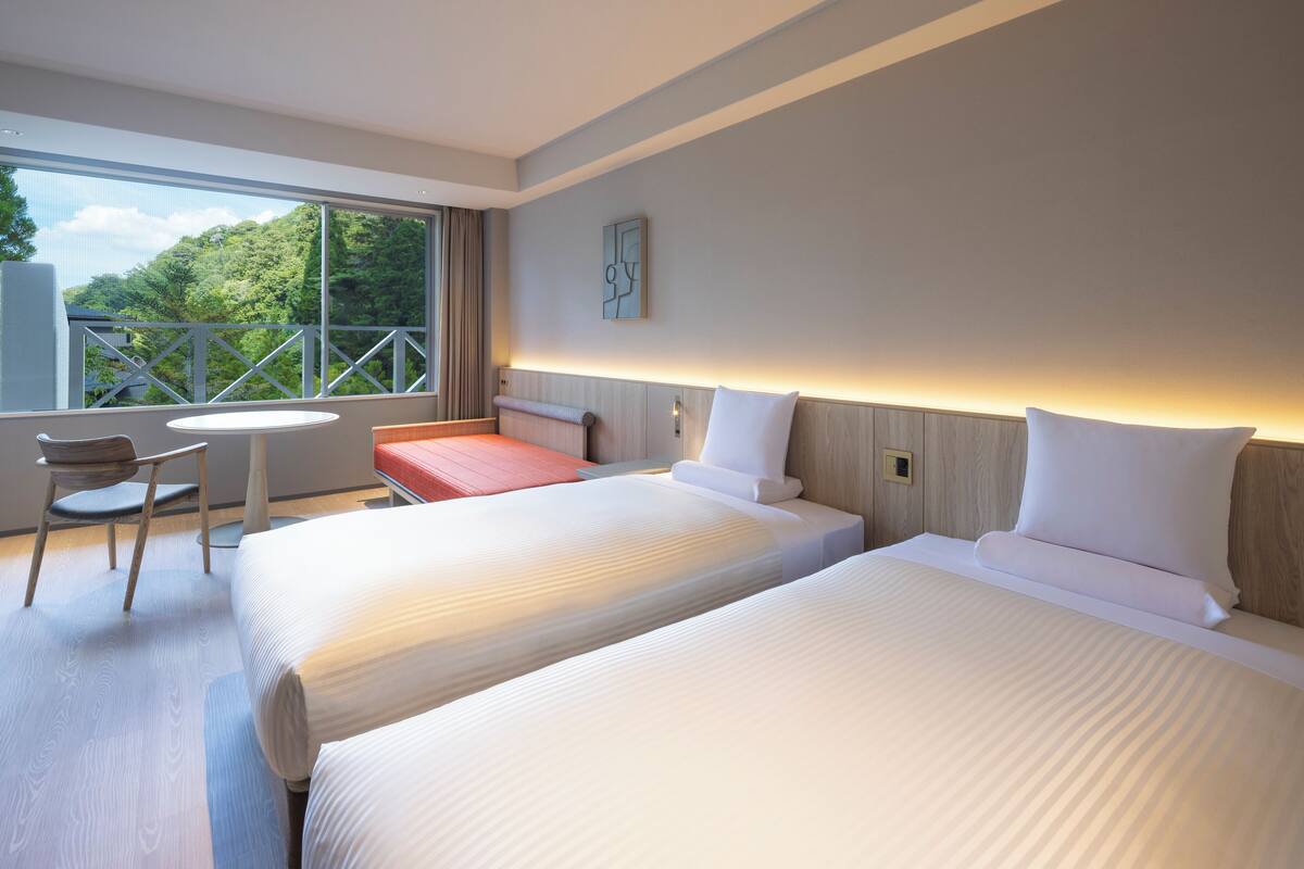 A hotel room with two beds, a small table, a chair, and a balcony with a view of greenery.