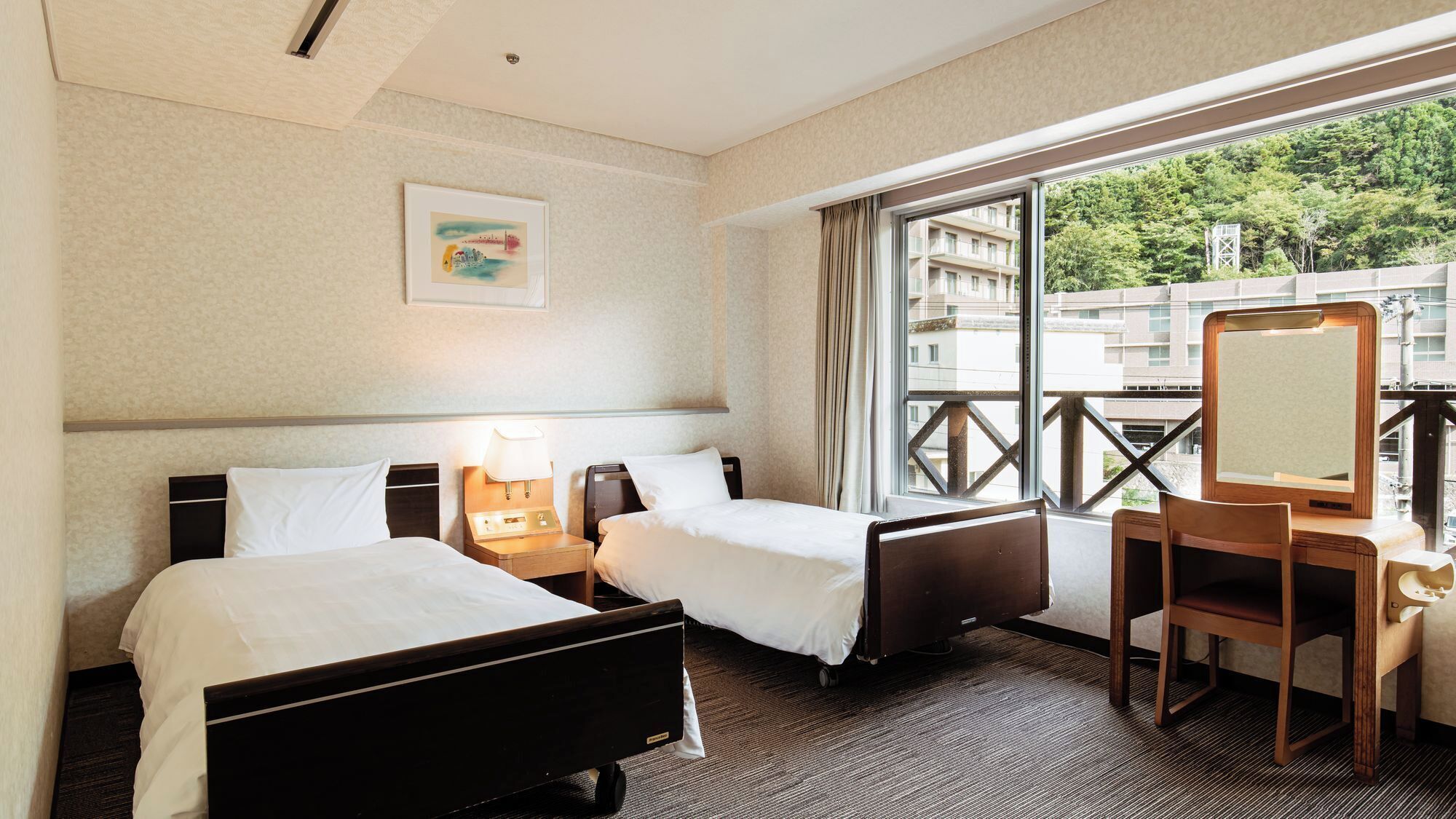 Une chambre d’hôtel avec deux lits, un bureau, une chaise, une fenêtre donnant sur des immeubles et un tableau au mur.