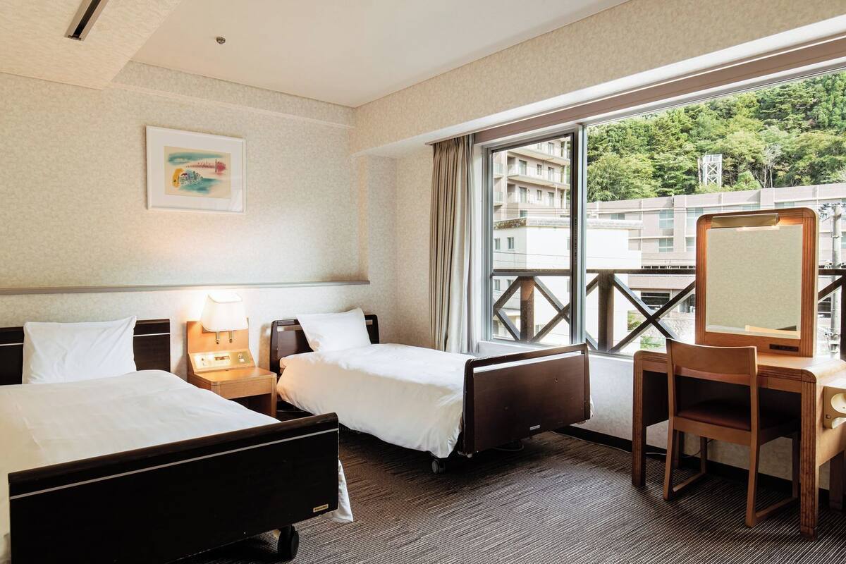 A hotel room with two beds, a desk, a chair, a window with a view of buildings, and a painting on the wall.