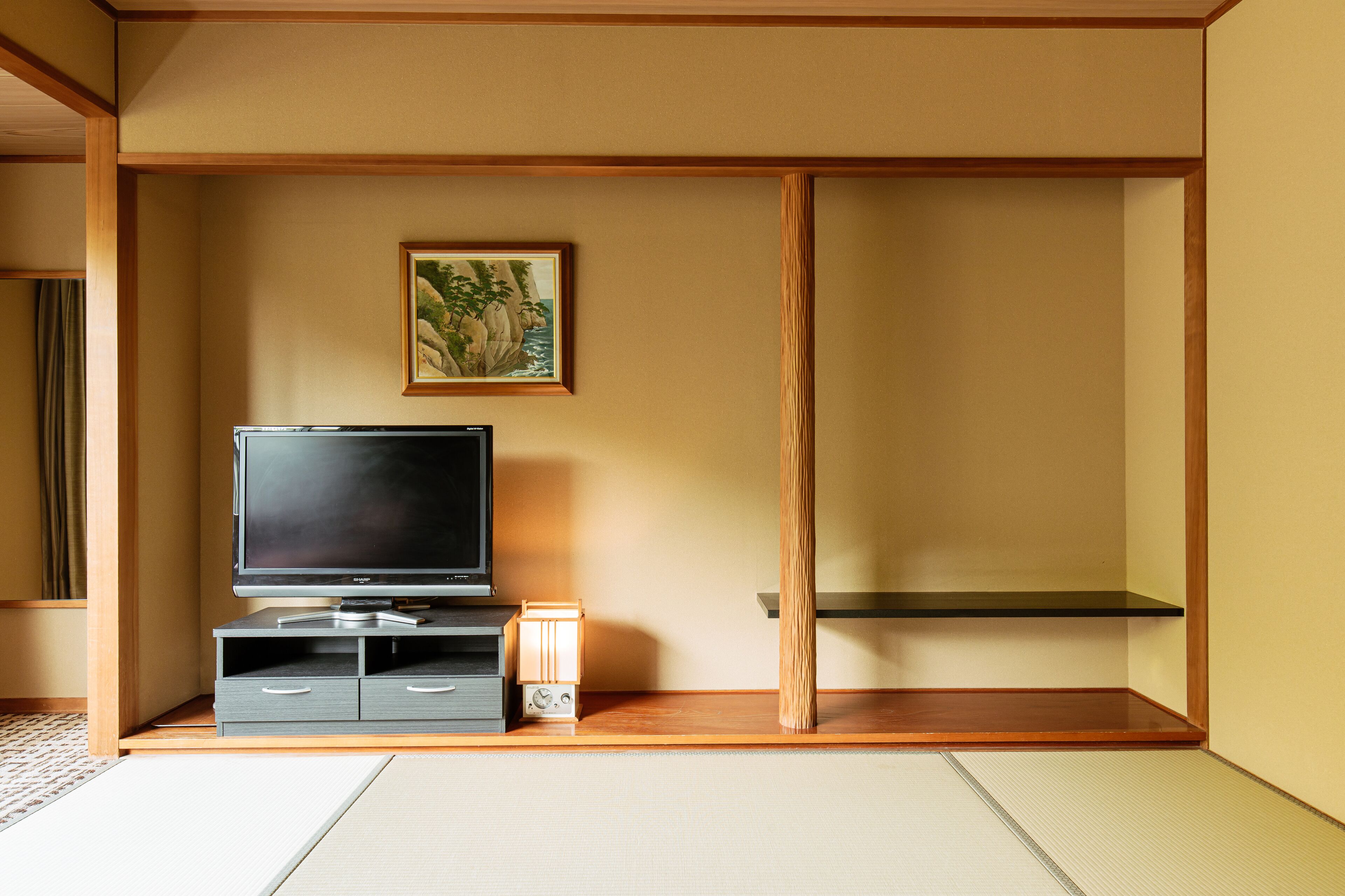 A minimalist room with tatami flooring, a television on a low stand, a small table with a lamp, and a framed picture on the wall.