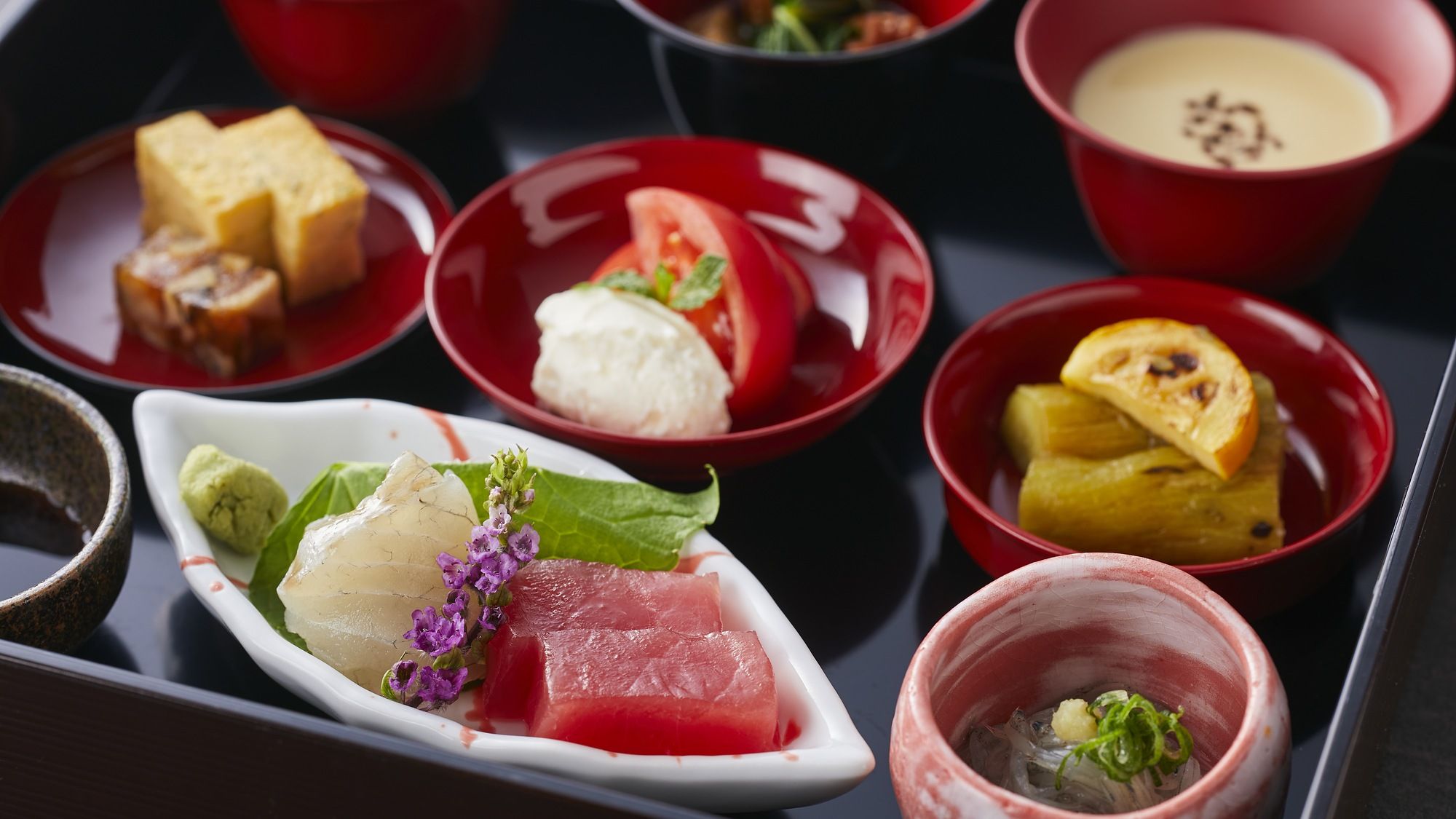 A variety of dishes arranged on a tray, including sashimi, a bowl of soup, and a plate with a slice of watermelon.