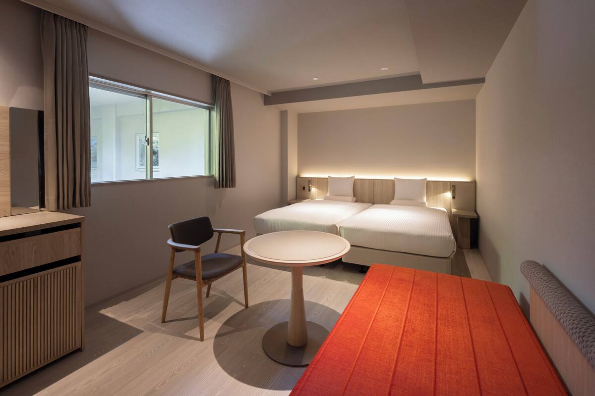 A modern hotel room with a bed, bedside tables, a desk, a chair, and a window with curtains.