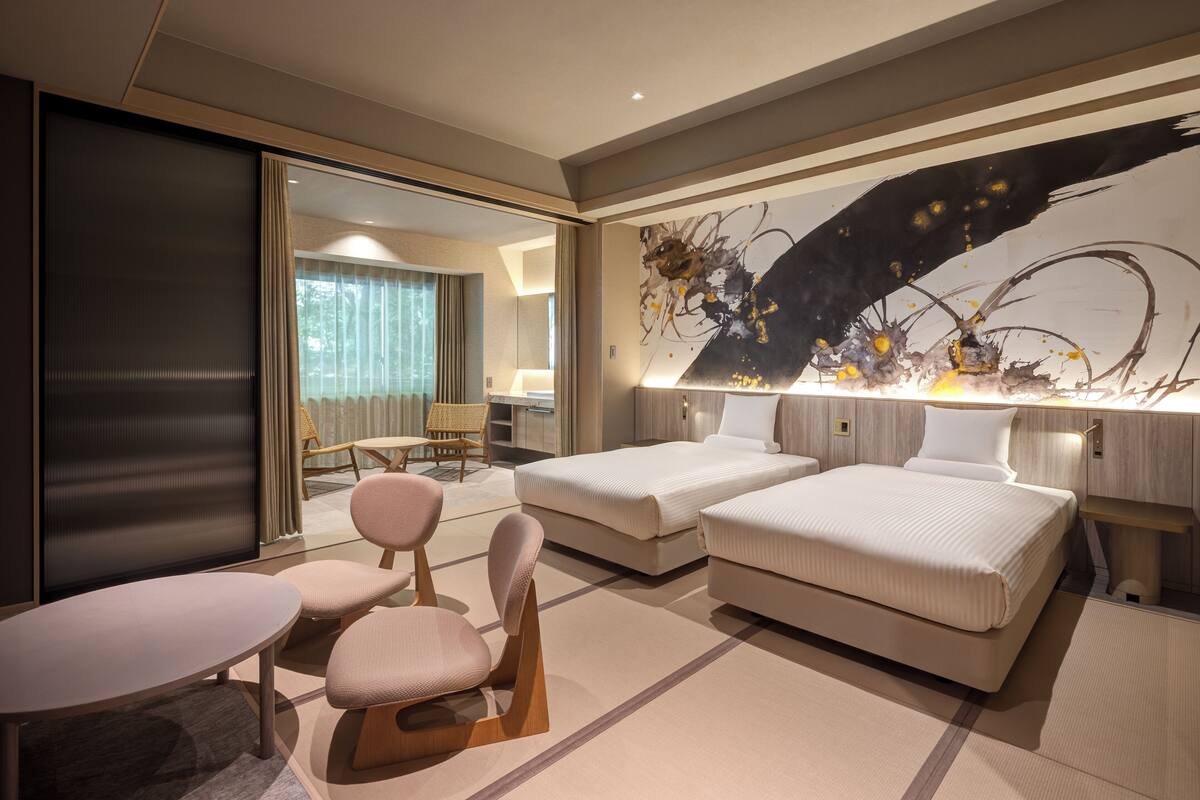 A modern hotel room with two beds, a sitting area with a table and chairs, a large abstract painting on the wall, and a view of the outdoors through a sliding door.