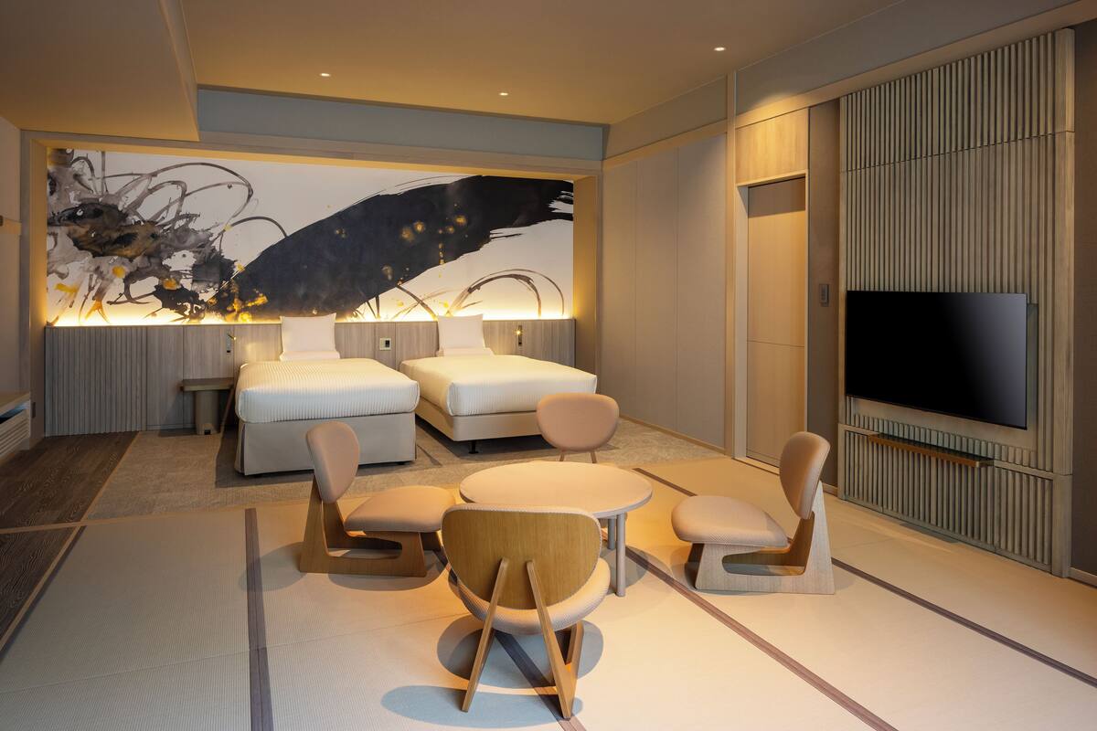 A modern hotel room with a large bed, a TV, a dining area with chairs, and a large abstract painting on the wall.