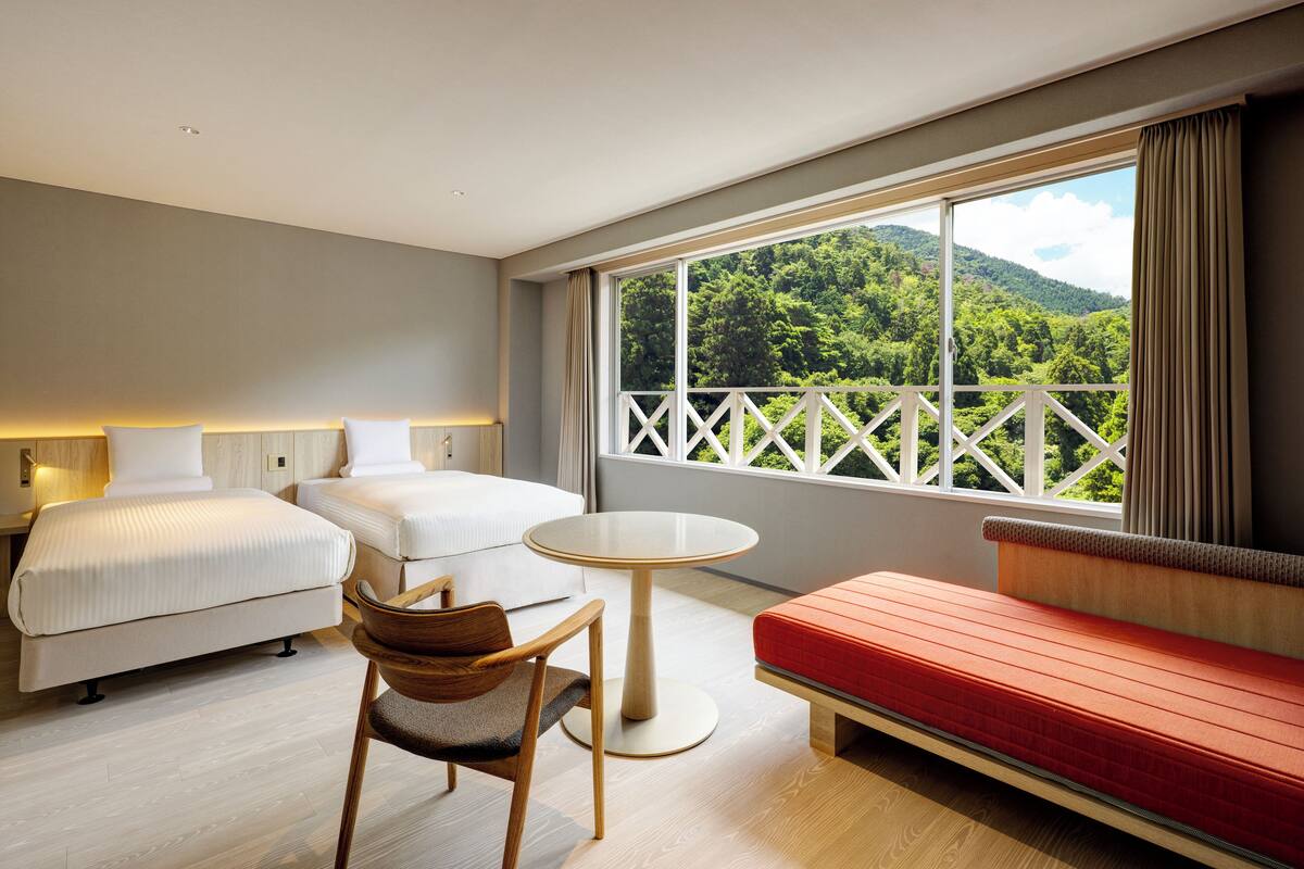 A hotel room with two beds, a round table, a red sofa, and a balcony with a view of green hills.