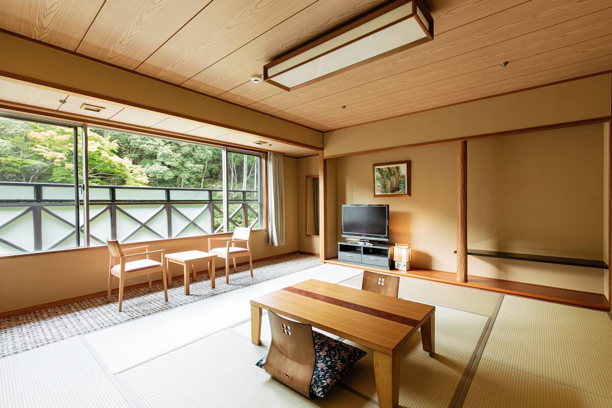 A traditional Japanese room with tatami flooring, a wooden table, chairs, a television, and a view of greenery outside.