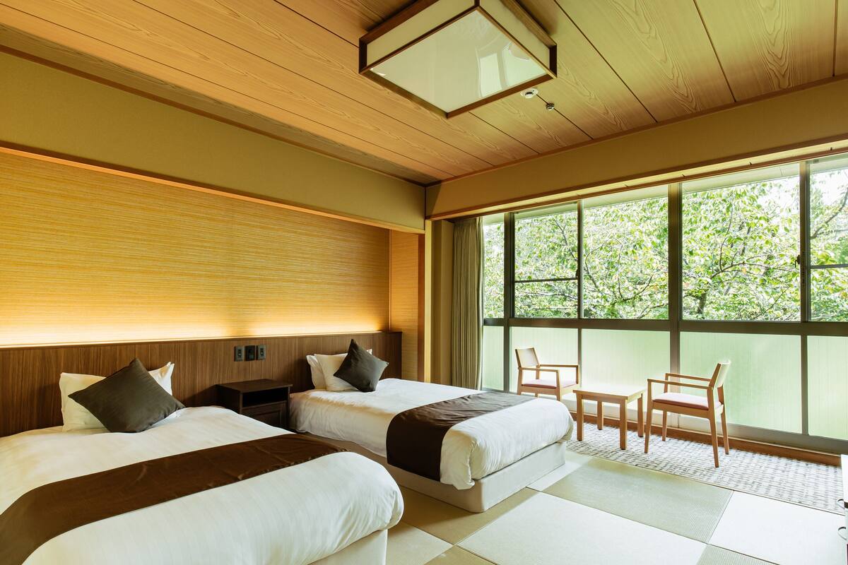 A hotel room with two beds, a wooden ceiling, large windows, and a view of trees.