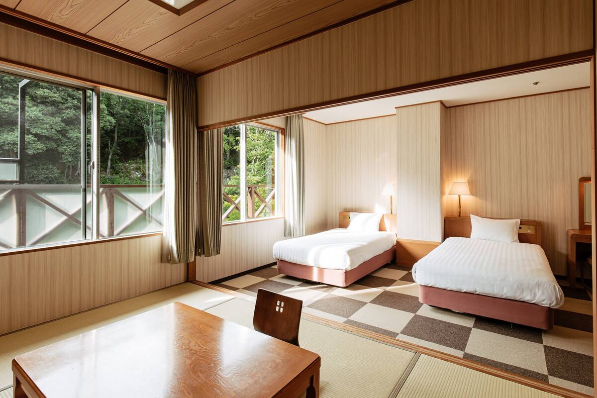 A hotel room with two beds, a wooden table, and large windows offering a view of greenery.