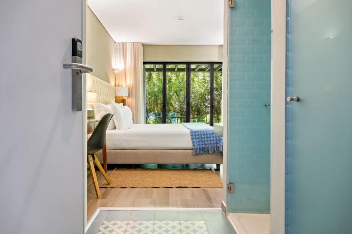 Een slaapkamer met een bed, nachtkastje en uitzicht op groen door een raam.