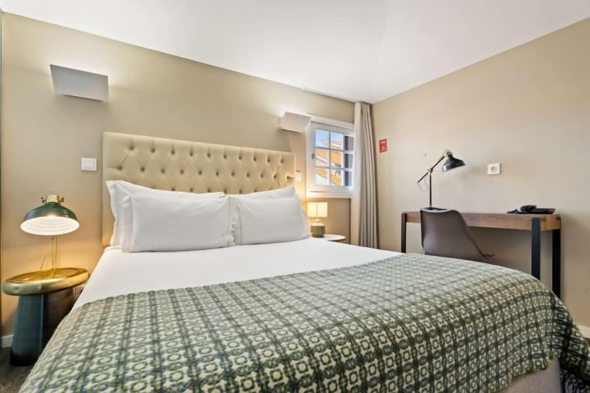 A hotel room with a large bed, a desk, a chair, a lamp, and a window.