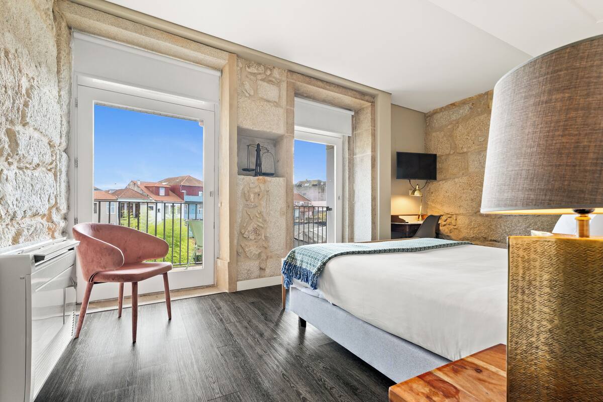 Ein Hotelzimmer mit Bett, Stuhl, Schreibtisch, Fernseher und einem Balkon mit Blick auf Gebäude.