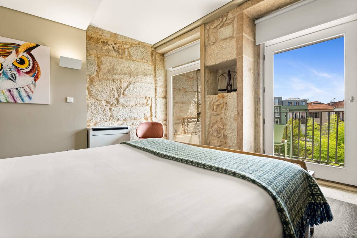 A bedroom with a large bed, a stone wall, a balcony with a view of buildings, and a painting of an owl.