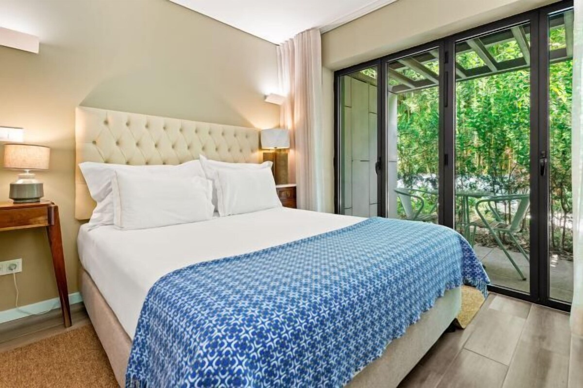 A hotel room with a large bed, a nightstand with a lamp, and a view of greenery through a sliding glass door.