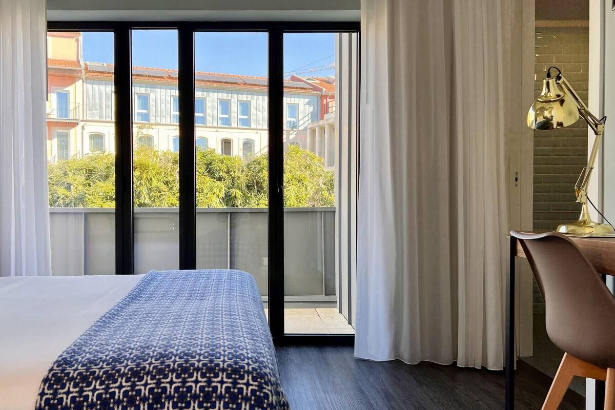 A hotel room with a bed, a desk, a chair, and a view of buildings and trees through large windows.