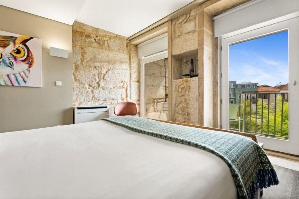 A hotel room with a large bed, a stone accent wall, a painting of an owl, and a balcony with a view of buildings.