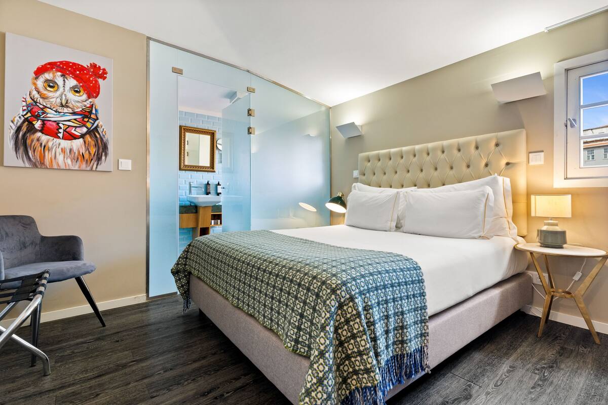 A hotel room with a large bed, a chair, a bedside table, and a painting on the wall.