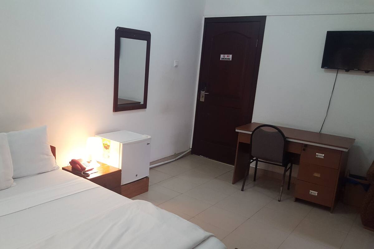 Una habitación de hotel con cama, escritorio, silla, armario y televisor en la pared.