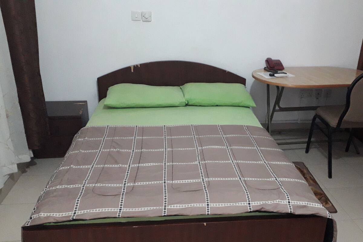 Una cama individual con un edredón a cuadros marrón y blanco y dos almohadas verdes. Junto a la cama hay una mesita con una silla, y un objeto rojo sobre la mesita.