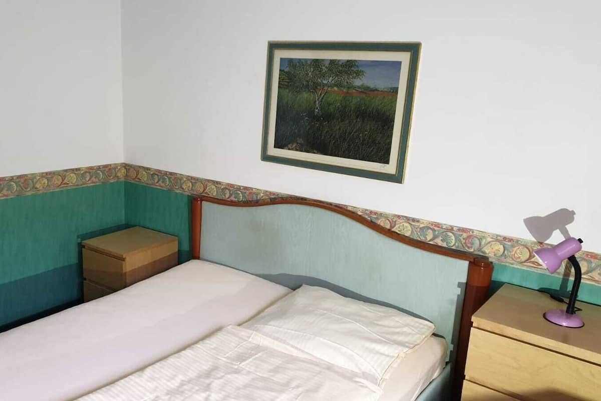 Uma cama de solteiro com cabeceira de madeira, uma mesinha de cabeceira com abajur, uma pequena mesa de apoio e um quadro na parede.