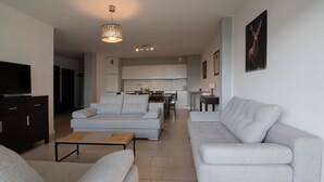 Ein modernes Wohnzimmer mit grauen Sofas, einem Couchtisch und einem Fernseher. Im Hintergrund ist eine Küche mit weißen Schränken und einem Essbereich zu sehen.
