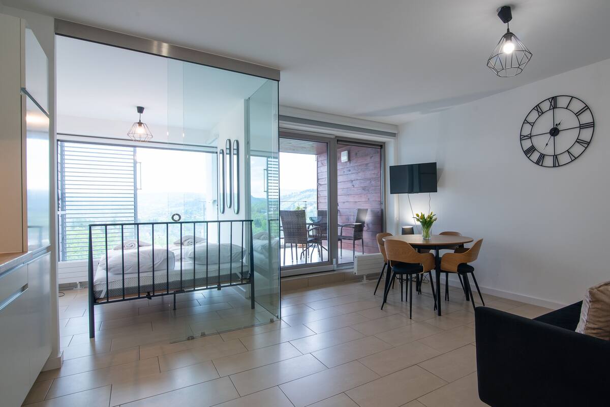 Un salon moderne avec une porte vitrée donnant sur un balcon, un coin repas avec une table et des chaises, un canapé noir et une horloge murale.