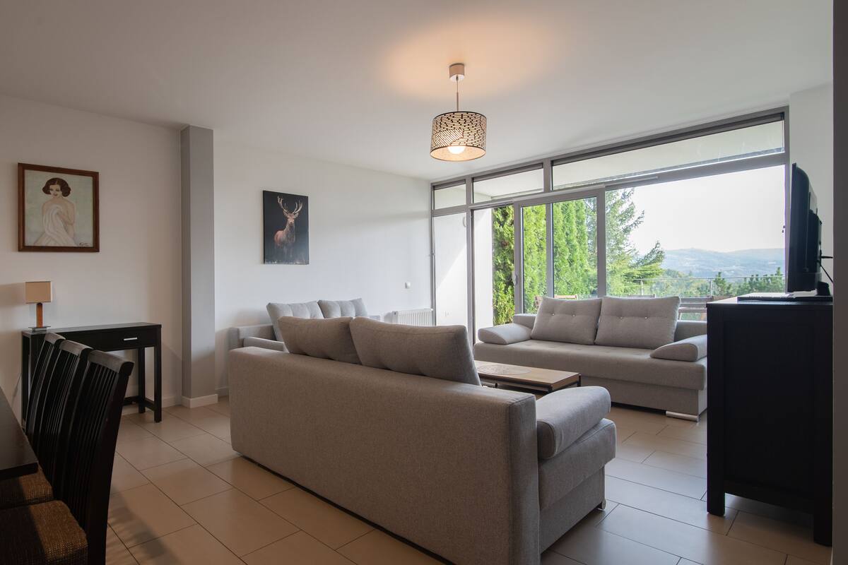 Un salon moderne avec un canapé, une table à manger et des chaises. Un tableau orne le mur, et une fenêtre offre une vue sur les arbres et les collines.