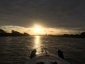 A boat's deck with a view of the sunset over a river, including distant buildings and trees.