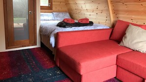 Una habitación acogedora con techo de madera, una cama con ropa blanca y almohadas rojas, un sofá rojo y una alfombra colorida.