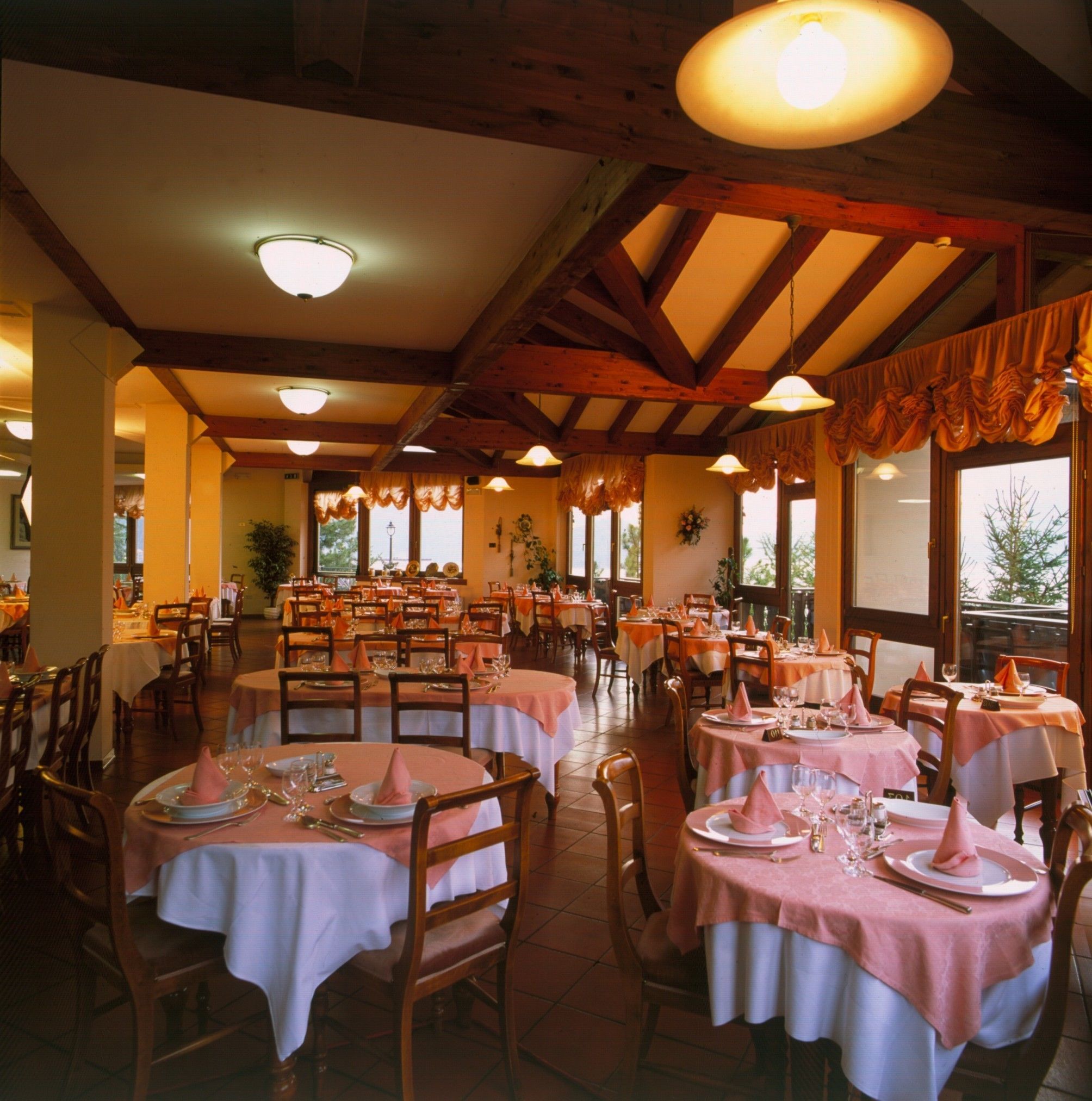 Un restaurante con varias mesas redondas cubiertas con manteles blancos y servilletas rosas, sillas de madera y un techo alto con vigas a la vista.