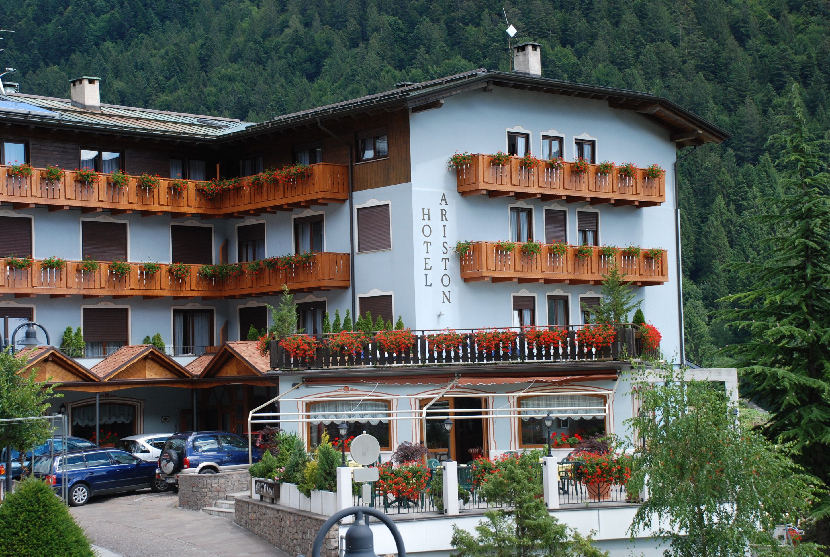 Un hotel con numerosi balconi decorati da fiori, una zona salotto esterna coperta e montagne sullo sfondo.