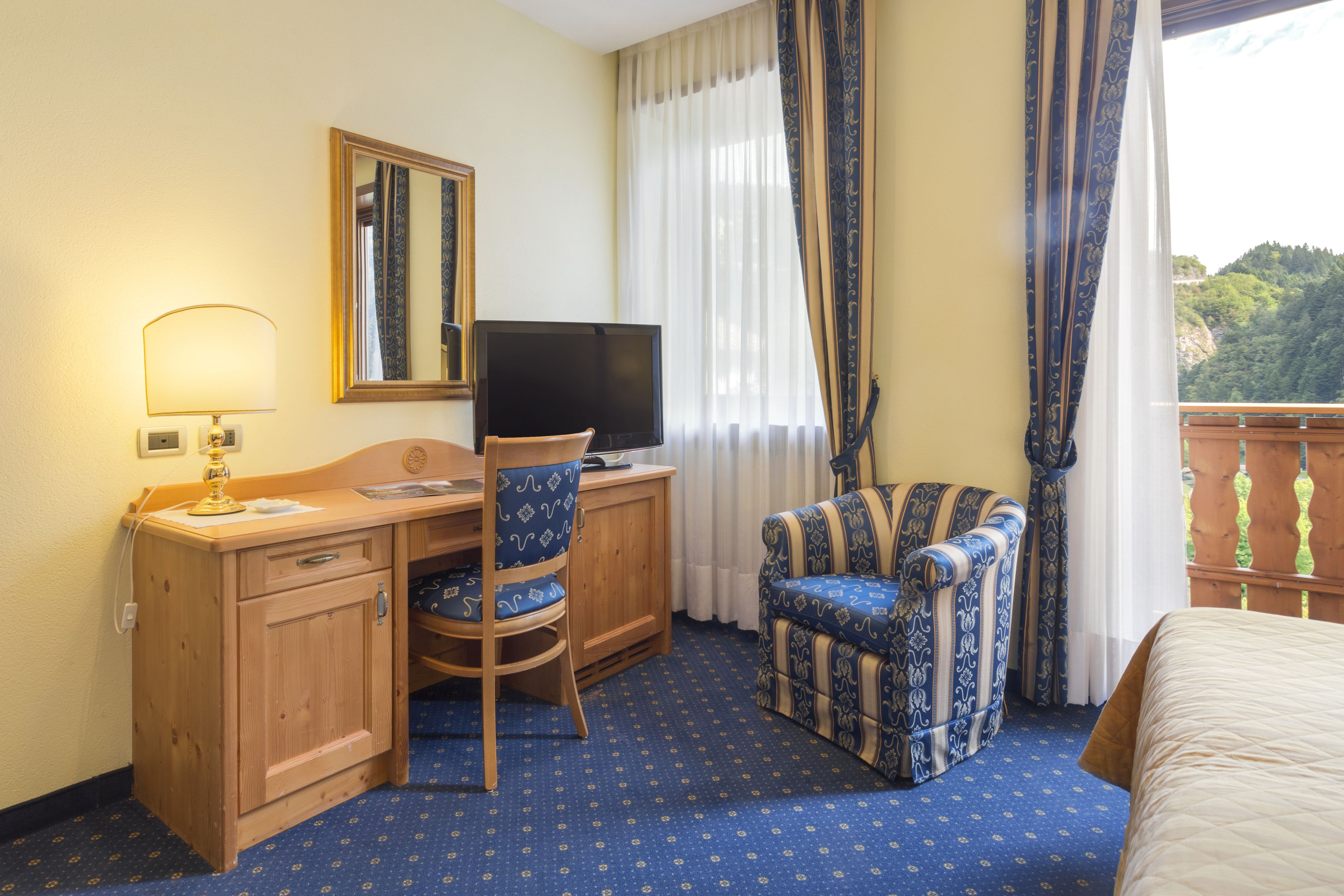 Habitación de hotel con escritorio de madera, una silla, televisor, cama y balcón con vista a vegetación.